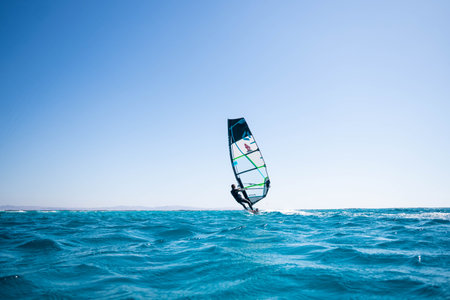 A rear view of a man windsurfing in splashes of turquoise water with blue sky in the backgroundの写真素材