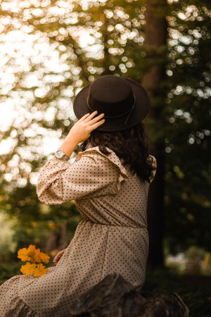 A vertical shot of a brunette female from back in a dress and elegant hat sitting in the forestの写真素材