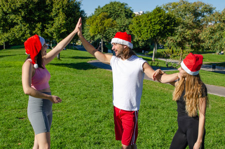 Picture of Happy Friends in Sportswear with Santa Claus Christmas Hats High-fiving their Hands while Smiling in a Park Backgroundの写真素材