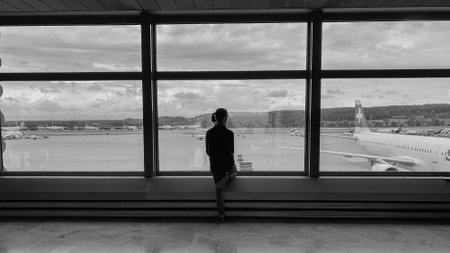 A grayscale of a young woman looking at an airplane from a glass windowの写真素材