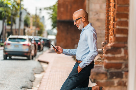 A bald Hispanic male using his smartphone while leaning against a brick wallの写真素材