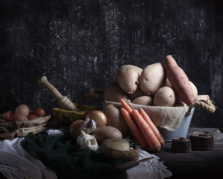 A metal bowl of potatoes with onions, heads of garlic, carrots and chicken eggs on a wooden surfaceの写真素材
