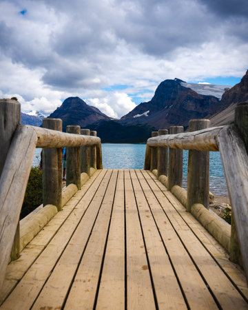 A pov of a wooden bridge leading to a lake with a mezmerising view of the mountains under a cloudy skyの写真素材