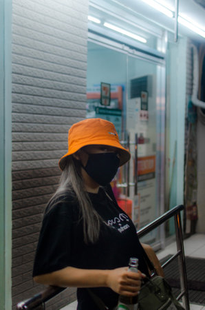 A woman in a hat and a mask holding a bottle and leaning on a fence in the streetの写真素材