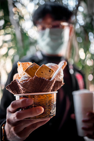 A vertical shot of a young male holding a paper bowl with ice cream and biscuit toppingsの写真素材