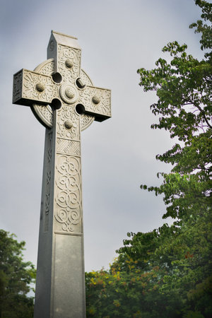 The Celtic stone cross in the park near green trees against a blue sky on a gloomの写真素材