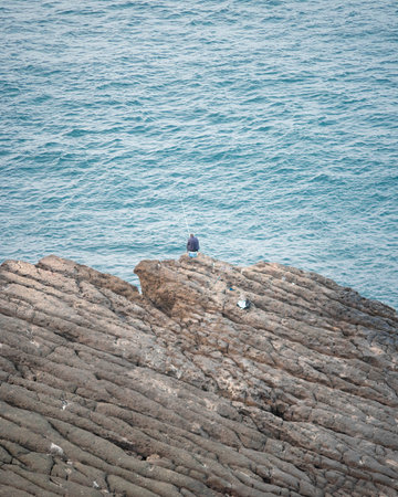 A vertical shot of a lone male fishing on a large cliffの写真素材
