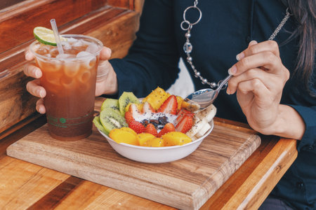 A woman eating a fruit salad with iced tea on a wooden tableの写真素材