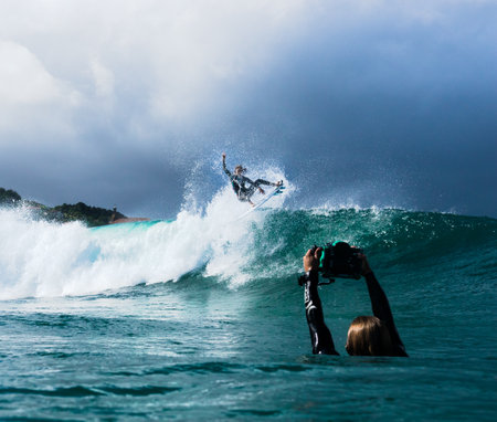 A surfer taking a picutre of his friend surfing the foamy wave of the oceanの写真素材