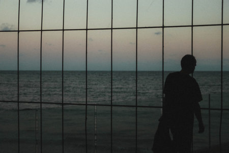 A rear view of a male behind large grids looking towards the peaceful lake water at sunsetの写真素材