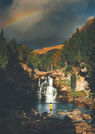 A glorious scenery of a rainbow in the sky above a waterfall flowing into the river in wild woodsの写真素材