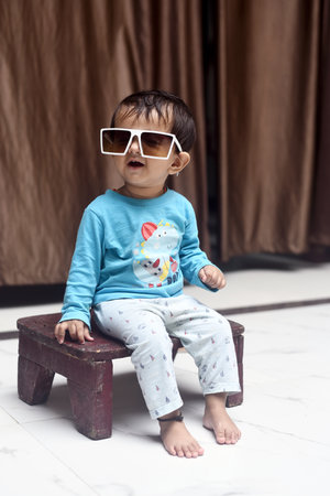 A vertical closeup of the cute baby girl with sunglasses sitting on a wooden chair.
