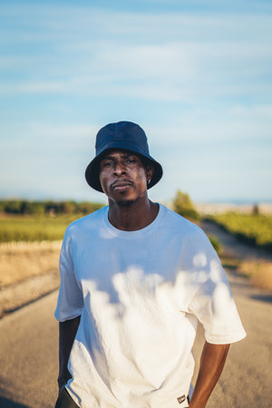 A portrait of a young black handsome man wearing a bucket hat walking along the highway at sunset