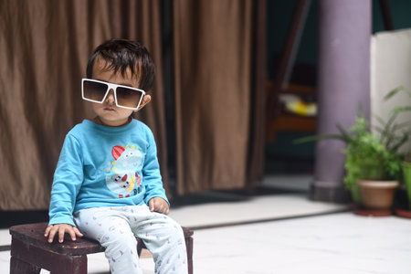 A closeup of the cute baby girl with sunglasses sitting on a wooden chair.