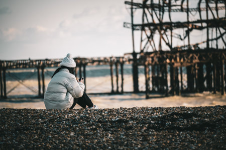A lonely woman wearing a coat and a hat and sitting on the beach with an old pier in the backgroundの写真素材