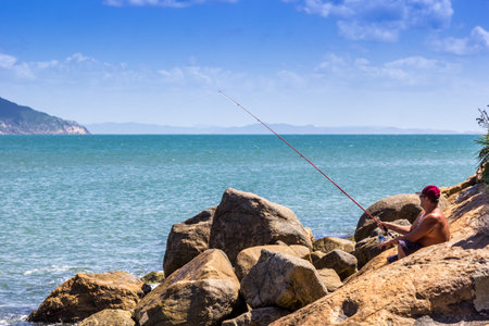 A beautiful view of a male sitting on a rocky beach fishing  using a fishing rod in the sunny seaの写真素材