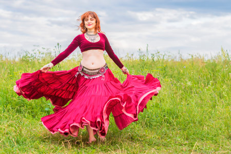 Young beautiful red-haired girl in a red dress enthusiastically dances a fascinating dance on a green lawn under the open blue sky.の写真素材