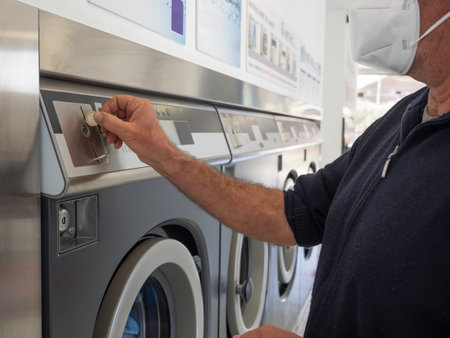 A man wearing a sanitary mask inserting a coin in a washing machine in a public laundromatの写真素材