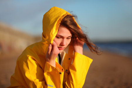 Young girl wearing a bright yellow raincoat, putting on a hood in windy weather on the beach.