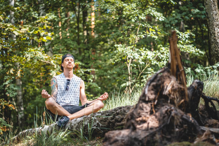 A young adult man with tattoos meditating in a forest on a sunny day in Berlin, Germanyの写真素材