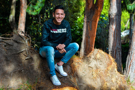 A young cheerful Colombian male sitting near the trees and smiling