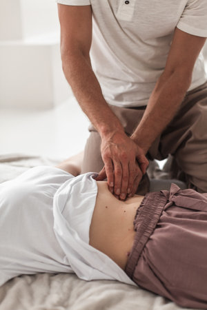 A professional young man massaging a woman's abdomen in a white cozy room during a manual therapy
