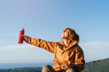 A Caucasian female hiker in an orange coat holding a water bottle and enjoying the warmth of the sunの写真素材