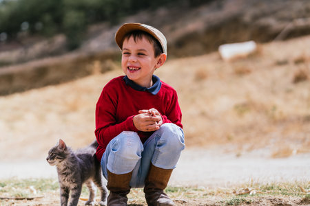 A Caucasian village boy wearing boots and a hat with a kitten on a farmの写真素材