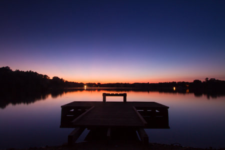 A wooden pier over the lake water surface with forest in the background against clear sky at sunsetの写真素材