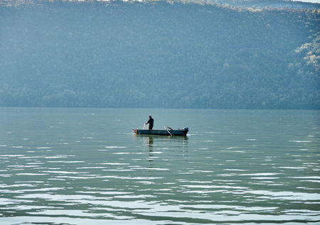 A fisherman on a fishing boat on a lake surrounded by hills under the sunlightの写真素材