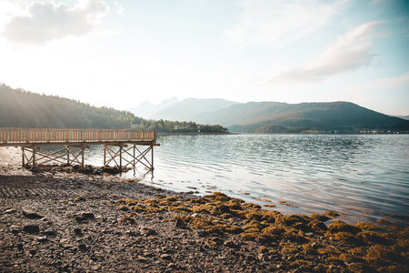 A beautiful view of wooden pier at lakeshore against mountains and blue cloudy sky in bright sunlightの写真素材