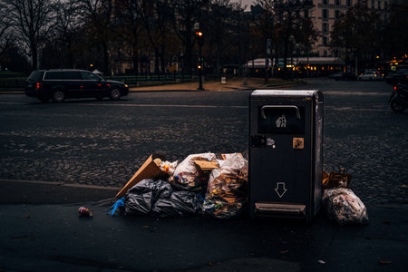 A trash bin with trash bags next to it on the sidewalk