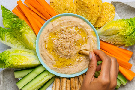 White bean hummus alongside cut vegetables. A woman's hand dipping the vegetable into the bowl....