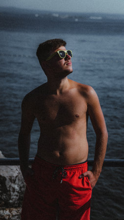 A portrait of a shirtless young British man wearing red shorts at the beach on a sunny day