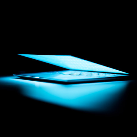 A closeup shot of a half-open laptop shining light on a dark tableの写真素材