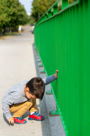 A vertical shot of a Caucasian young boy squatted holding a green metallic fenceの写真素材