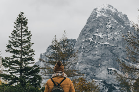 A Caucasian female hiker from behind looking straight at pine trees with a background of sky and mountains covered with snow in Sloveniaの写真素材