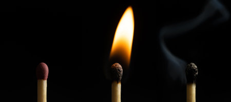 A closeup of three matches of an extinguished match next to a burning one isolated on the black...