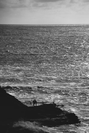 A vertical grayscale shot of fishermen on a cliff near a seaの写真素材