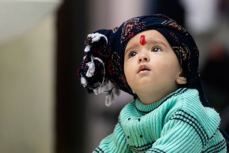 A South Asian baby girl wearing green winter sweater and red turban standing on a toy horse