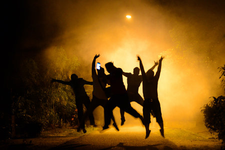 The silhouettes of a group of people jumping up against a brown light at nightの写真素材