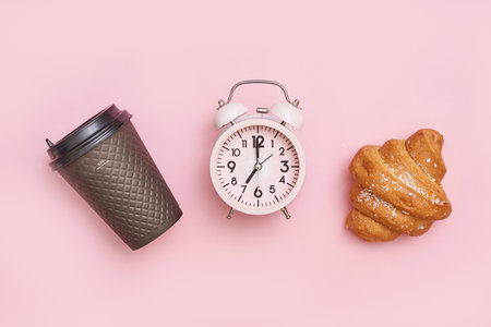 The vintage alarm clock with a cup of coffee and a fresh croissant on the pink surfaceの写真素材