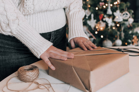 Midsection of woman wearing cozy thick sweater, wrapping christmas present in kraft paper. Christmas tree with bokeh lights in background.の写真素材