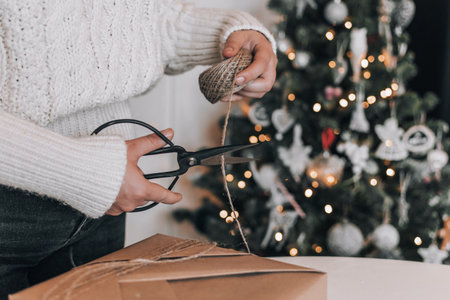 Midsection of woman wearing cozy thick sweater, wrapping christmas present in kraft paper. Christmas...