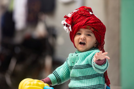 A South Asian baby girl wearing green winter sweater and red turban standing on a toy horseの写真素材