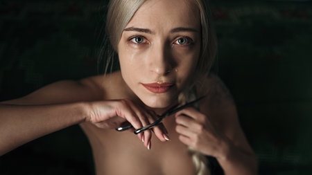 A beautiful blond Caucasian girl with tears in the eyes cutting her hair with the scissorsの写真素材