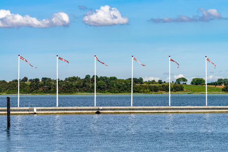 A body of water and a pier with six flags of Denmark waving on a stick against a light blue sky with...