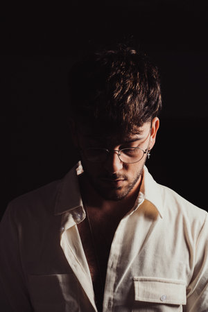 A portrait of a young sexy Hispanic man with glasses and beard wearing a white shirt, black...