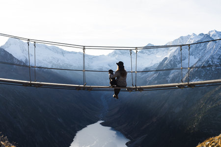 Girl traveler with her dog in the mountains. A young girl sit on a suspension bridge on a background...
