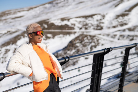 An African American woman leaning on a fence and wearing a winter jacket and glasses.